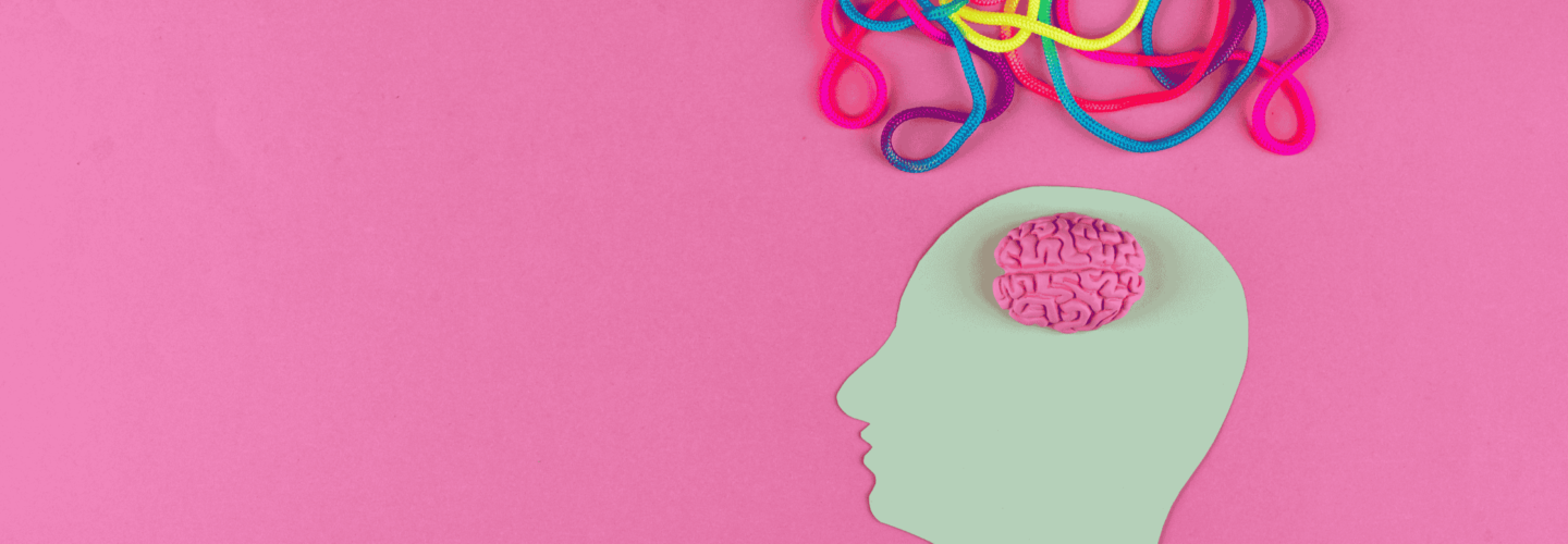 A minimalist and conceptual art piece featuring a pastel green paper cutout of a human head in profile on a pink background. A small, three-dimensional pink brain is placed inside the head cutout. Above the head, colorful tangled cords in shades of pink, blue, yellow, and purple extend outward, symbolising complex thoughts or neurodiversity.