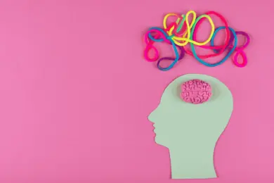 A minimalist and conceptual art piece featuring a pastel green paper cutout of a human head in profile on a pink background. A small, three-dimensional pink brain is placed inside the head cutout. Above the head, colorful tangled cords in shades of pink, blue, yellow, and purple extend outward, symbolising complex thoughts or neurodiversity.